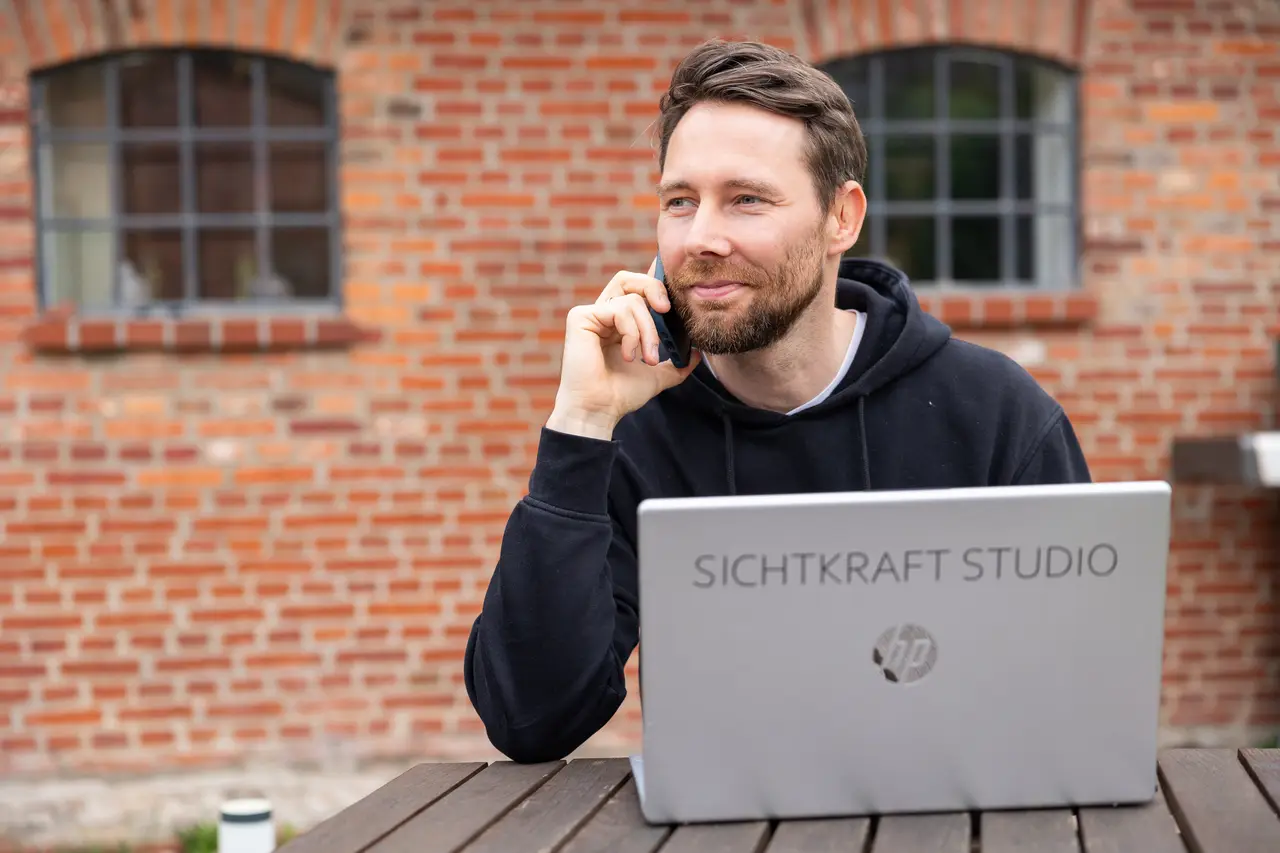 Dirk Grüner telefoniert vor einer Backsteinwand, auf dem Laptop-Deckel das Logo von Sichtkraft Studio