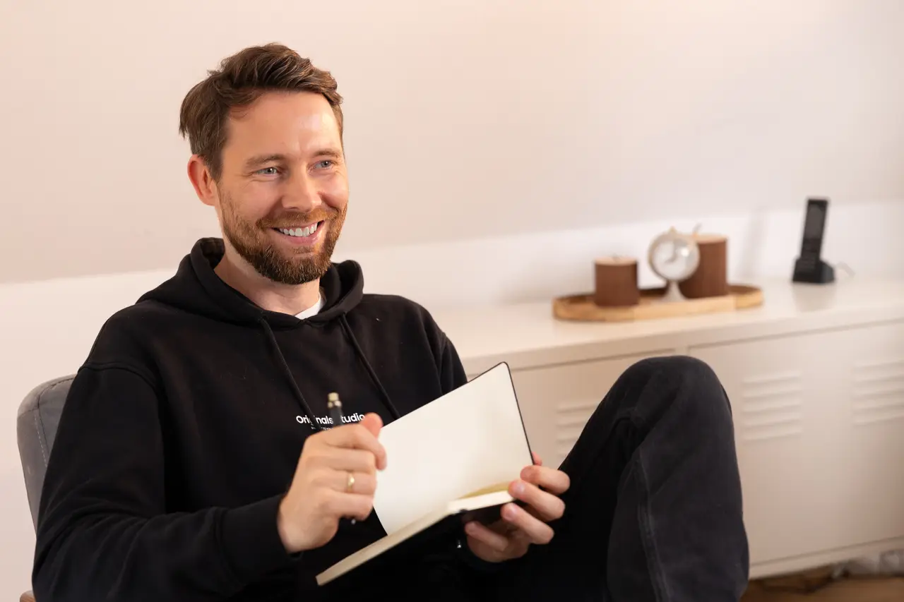 Dirk Grüner von Sichtkraft Studio sitzt im Büro auf einem Stuhl, lächelt in die Kamera und hält Notizblock und Kugelschreiber in der Hand