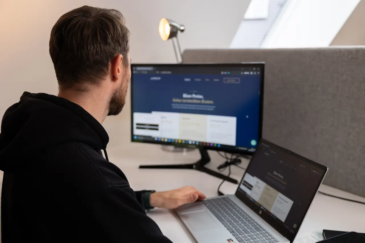 Dirk Grüner von Sichtkraft Studio sitzt am Schreibtisch und arbeitet an einer Webseite, auf Monitor und Laptop ist der Entwurf zu sehen