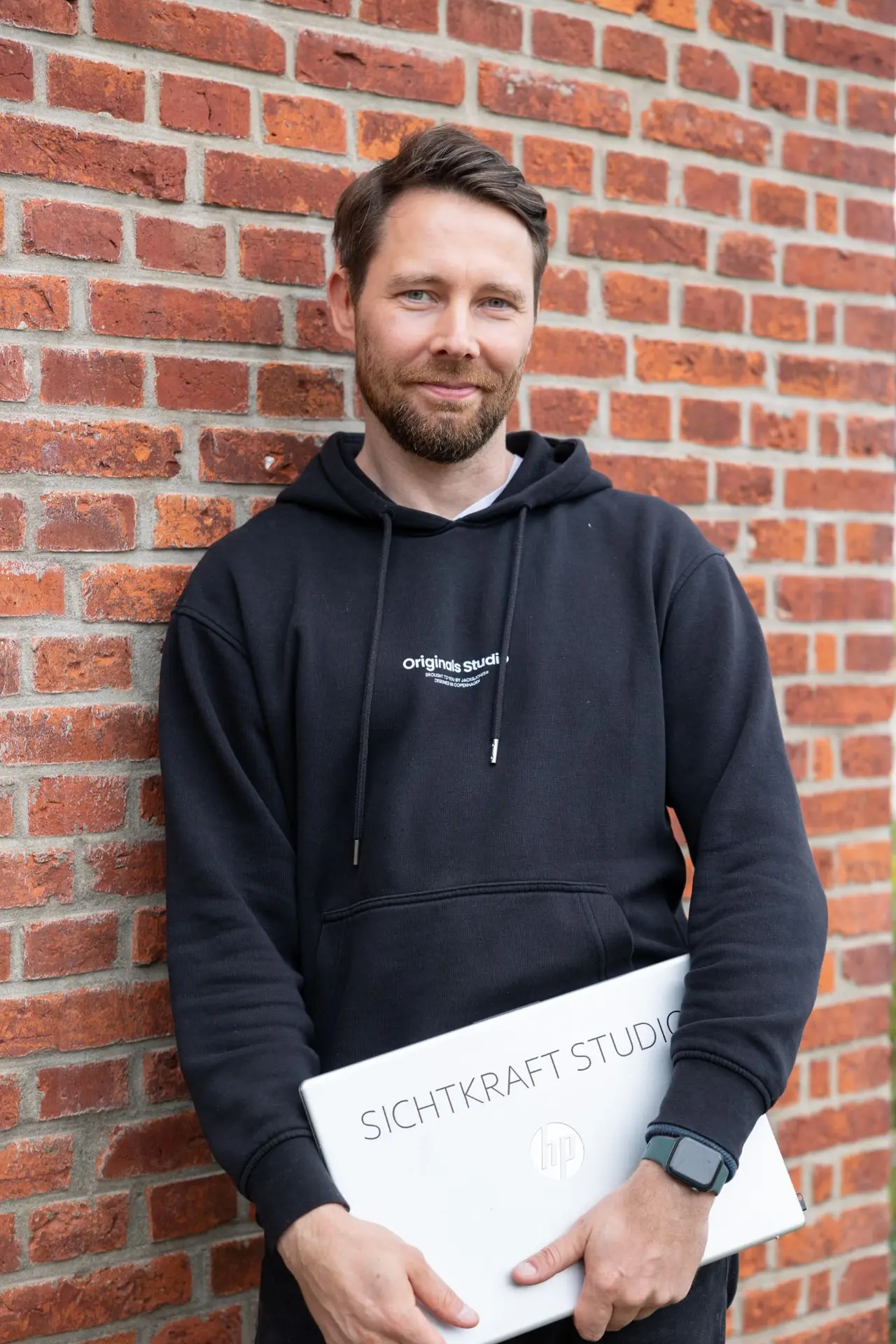 Dirk Grüner steht vor einer Backsteinwand und hält einen Laptop mit dem Sichtkraft-Studio-Aufkleber