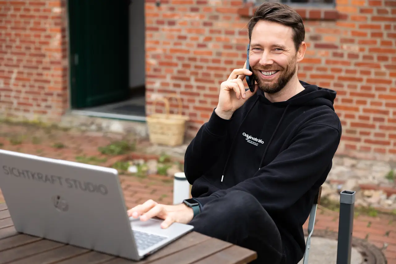 Dirk Grüner lacht am Telefon vor einer Backsteinwand, während er am Laptop mit der Aufschrift Sichtkraft Studio arbeitet