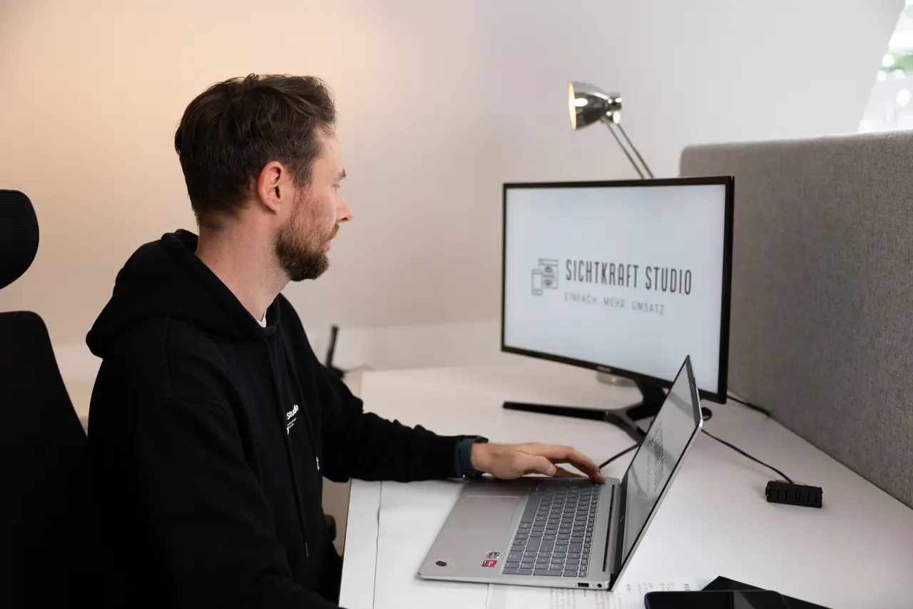 Dirk Grüner sitzt seitlich am Schreibtisch vor Laptop und Monitor, auf dem Monitor ist das Sichtkraft-Studio-Logo zu sehen
